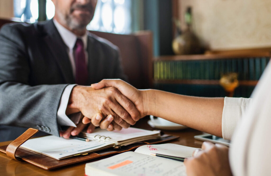A man wearing a suit and sitting at a wooden desk is building effective workplace relationships with his colleague who is just out of the picture — they are shaking hands.